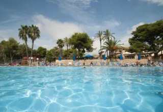 Large outdoor swimming pool with sun loungers, palm trees, and blue umbrellas at Vilanova Park, Catalonia, Spain.