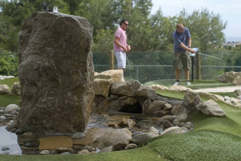Deux hommes jouent au mini-golf près d’un rocher et d’un ruisseau au Vilanova Park Campsite, Catalogne, Espagne.