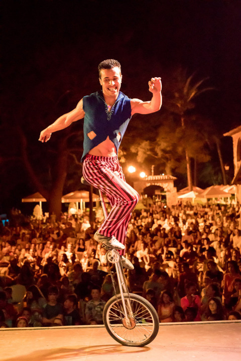 A performer rides a unicycle on stage in front of a large crowd at Vilanova Park Campsite in Catalonia, Spain.