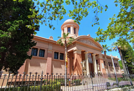 Edificio histórico en Vilanova i la Geltrú, Cataluña, España, rodeado de árboles y una verja en un día soleado.