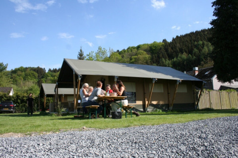 Familie nyder picnic foran telt på Camping Village Sy, feriepark i Liège, Belgien, omgivet af natur.