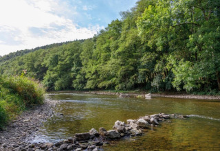 Scenic river with rocky shore and lush trees at Camping Village Sy, a holiday park in Liège, Belgium.