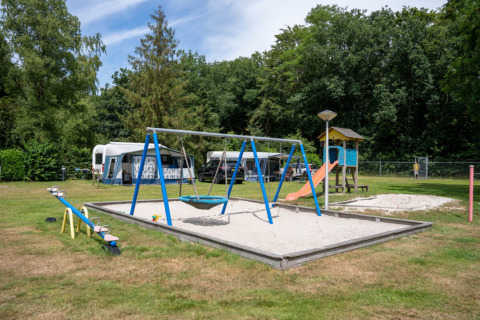 Legeplads med gynger, vippe og rutsjebane foran campingvogne på Camping de Zeven Heuveltjes i Drenthe.