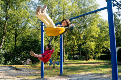 Dos niños juegan en columpios de un parque soleado en Camping de Zeven Heuveltjes, Drenthe, Países Bajos.