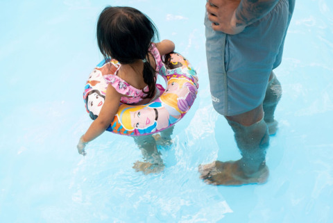 Lille pige med badering og voksen i shorts står i lavt vand i en swimmingpool på en campingplads.