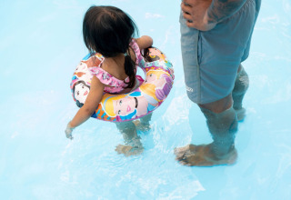 Lille pige med badering og voksen i shorts står i lavt vand i en swimmingpool på en campingplads.