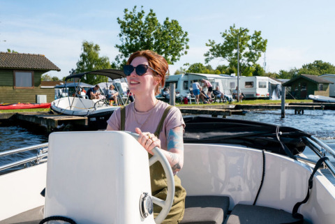 Frau steuert ein Boot bei Camping Tussen de Diepen in Overijssel, Niederlande, mit Wohnwagen im Hintergrund.