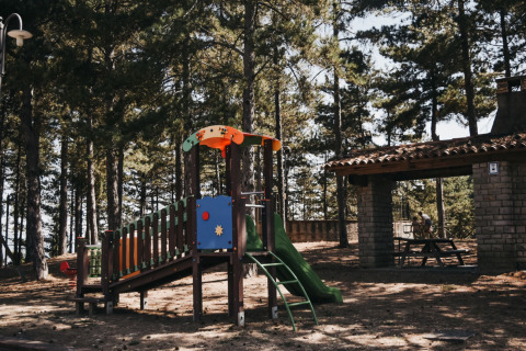 Parque infantil en Camping Luna del Monte, Umbría, Italia, rodeado de pinos, con tobogán y merendero.