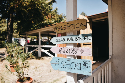 Træskilte viser vej til reception, poolbar, gelato og pool ved Camping Luna del Monte i Umbrien, Italien.