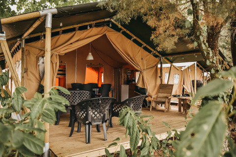 Tienda safari con terraza de madera, sillas de jardín negras y vegetación alrededor, ambiente acogedor.