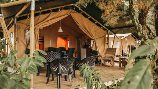 Tienda safari con terraza de madera, sillas de jardín negras y vegetación alrededor, ambiente acogedor.