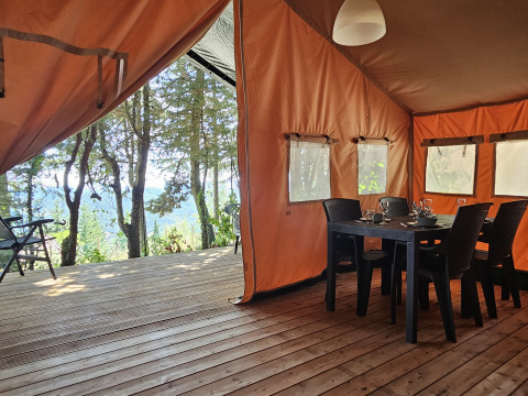 Tienda safari en Camping Luna del Monte, Italia, con terraza de madera, mesa de comedor y vistas al bosque.