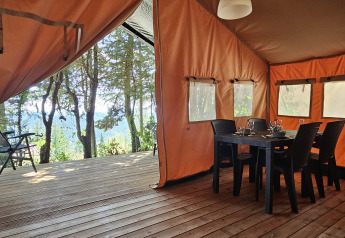 Tienda safari en Camping Luna del Monte, Italia, con terraza de madera, mesa de comedor y vistas al bosque.