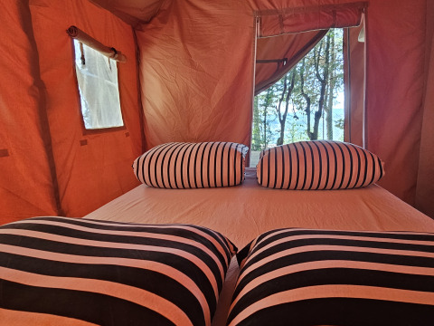 Interior de una tienda safari en Camping Luna del Monte, Italia, con almohadas a rayas y vista a los árboles.