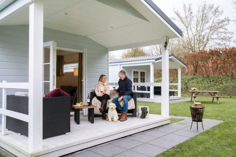 Couple relaxing with their dog on the porch of a Veranda Chalet at Camping Sevink Molen, Netherlands.
