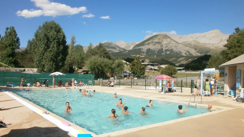 Piscina al aire libre en Camping Lou Passavous, personas nadando y vistas a las montañas bajo el sol.