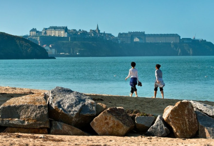 To personer går på stranden med udsigt over havet og de historiske bygninger ved Longueville i Normandiet.