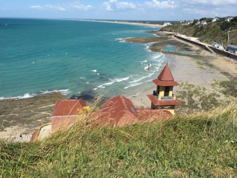 View of the beach and blue sea near Longueville, Normandy, France, with a coastal building in the foreground.