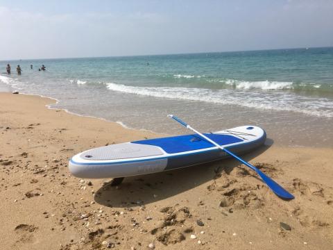 A paddleboard with paddle rests on a sandy beach near the sea, with people swimming in the background.