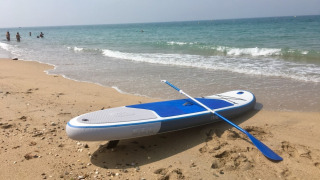 En paddleboard med åre hviler på en sandstrand ved havet, med mennesker badende i baggrunden.