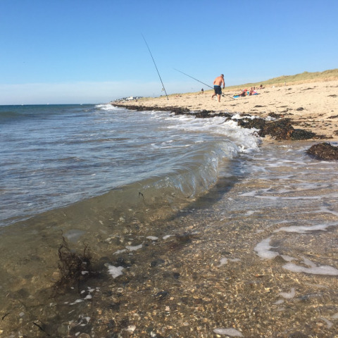 Strandbillede fra Camping O2 i Normandiet, Frankrig, med fiskestænger, bølger og badegæster.