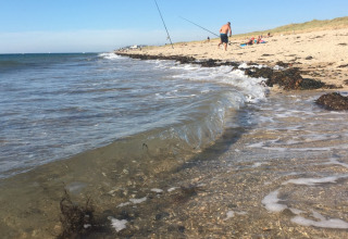 Strandbillede fra Camping O2 i Normandiet, Frankrig, med fiskestænger, bølger og badegæster.