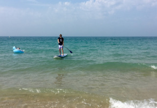 A person stands on a paddleboard and another relaxes on an inflatable lounger in the sea at a Normandy beach.
