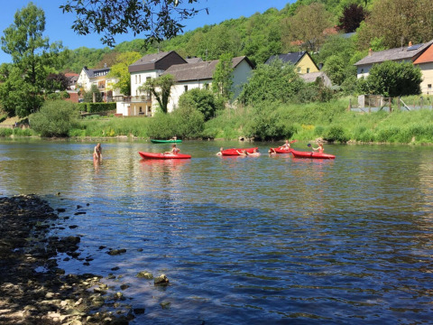 Children paddle red kayaks on a river at Camping du Rivage, with houses and trees in Diekirch, Luxembourg.