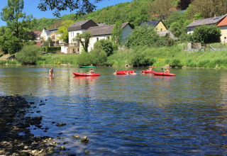 Children paddle red kayaks on a river at Camping du Rivage, with houses and trees in Diekirch, Luxembourg.