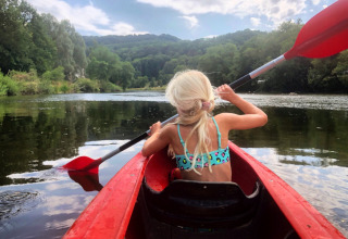 Blond meisje in badpak peddelt in een rode kajak op een rivier bij heuvels bij Camping du Rivage.