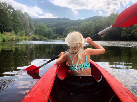 Blonde girl in swimsuit paddling a red kayak on a river, surrounded by hills at Camping du Rivage, Luxembourg.