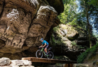 Cyclist rides over a small wooden bridge beneath large rocks at Camping du Rivage in Diekirch, Luxembourg.