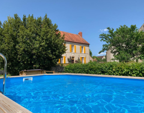 Piscine bleue devant une maison jaune aux volets orange en Auvergne-Rhône-Alpes, France, sous ciel ensoleillé.