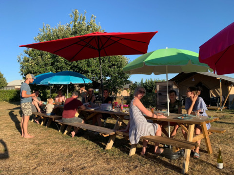 Menschen genießen ein Picknick an langen Holztischen mit bunten Sonnenschirmen auf einem Campingplatz.