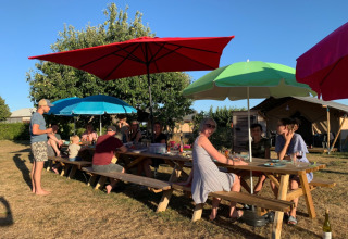Group of people at long wood tables with colorful umbrellas enjoying a sunny day at a French holiday park.