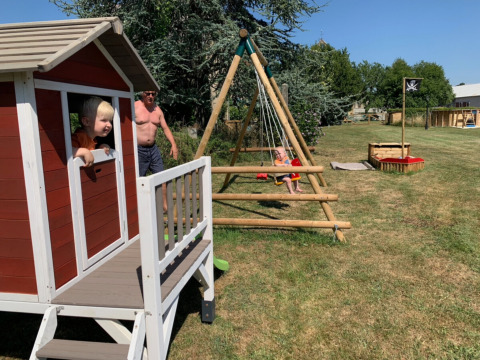 Des enfants jouent dehors dans une maisonnette, sur des balançoires et dans un bac à sable au camping.