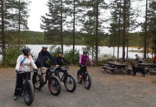 Cuatro personas en bicicletas fatbike junto al lago y rodeados de árboles en Camping Yttermalungs, Dalarna, Suecia.
