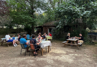 Folk nyder et måltid udendørs ved Domaine La Sauzette feriested, omgivet af træer i Occitanie, Frankrig.