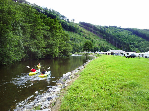 Dos personas en kayak en un río tranquilo junto a Camping la Roche, rodeados de colinas verdes y alojamientos.