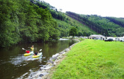 Two people kayaking on a calm river near Camping la Roche, surrounded by green hills and holiday homes.