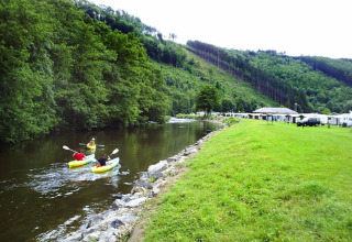 Two people kayaking on a calm river near Camping la Roche, surrounded by green hills and holiday homes.