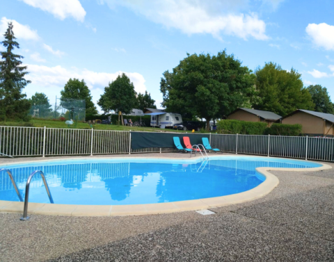 Piscine extérieure avec transats et clôture, entourée d’arbres, de caravanes et de chalets au Camping du Lac.