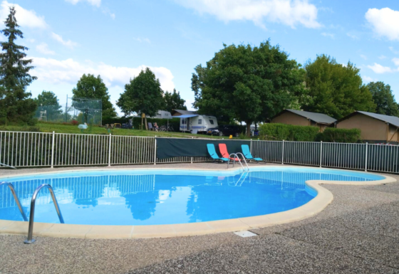 Piscina al aire libre con tumbonas y vallas, rodeada de árboles, caravanas y cabañas en Camping du Lac.