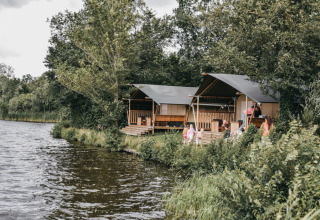 Cabanes au bord du lac au Camping de Kleine Wielen, Friesland, Pays-Bas, entourées d’arbres verts.