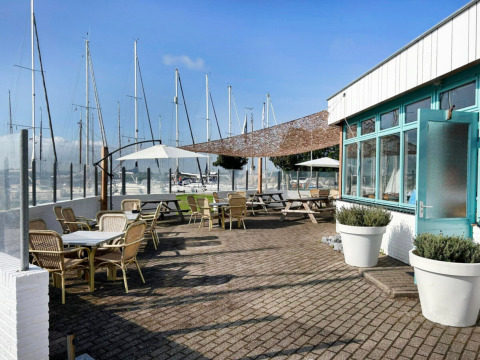 Außenterrasse mit Sitzplätzen bei Minicamping Marina Warns, Friesland, mit Blick auf Boote im Hafen.