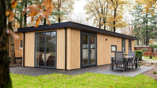 Moderne træhytte kaldet Module Wood med store vinduer og terrasse, omgivet af grønne træer om efteråret.