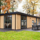 Moderne træhytte kaldet Module Wood med store vinduer og terrasse, omgivet af grønne træer om efteråret.