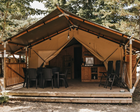 Tente safari avec sanitaires, terrasse en bois, table à manger et chaises dans un cadre boisé.