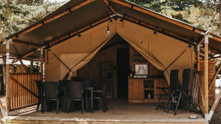 Tenda safari con servizi igienici, veranda in legno, tavolo e sedie immersi nella natura.