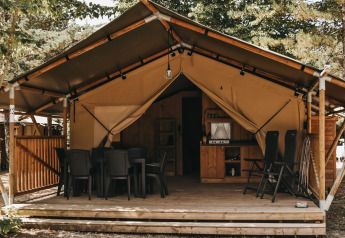 Tenda safari con servizi igienici, veranda in legno, tavolo e sedie immersi nella natura.